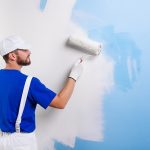 Back view of handsome young painter in white dungarees, blue t-shirt, cap and gloves painting a wall with paint roller.
